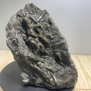 Peut inclure: Un spécimen minéral brut composé d'une matrice rocheuse gris foncé texturée, incrustée de nombreux cristaux de tourmaline verte translucides et allongés. La pierre présente une surface irrégulière avec des structures cristallines visibles.