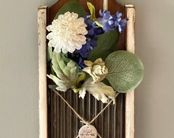 May include: A rustic wall hanging featuring a repurposed vintage grater. It's adorned with a green burlap bow, black and white checkered ribbon, and a floral arrangement of white and blue flowers. A small tag reads "Wild Free".
