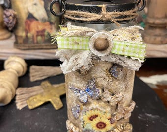 May include: A decorative glass jar featuring a textured, beige finish adorned with butterfly and sunflower motifs. The jar is topped with a black metal lid, a twine wrap, and a layered bow made of frayed fabric and green gingham ribbon with a central wooden button.
