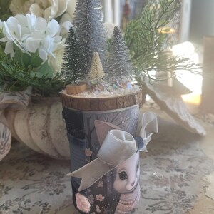 May include: A decorative jar with a winter scene, featuring miniature trees on a wooden base. The jar is decorated with a bunny illustration, a bow, and a winter forest design. A unique holiday decoration.