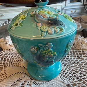 May include: A turquoise ceramic lidded bowl featuring raised relief designs of peacocks and floral patterns. The bowl has a pedestal base and a matching lid topped with a pointed black finial. The surface has a glossy finish with green and blue accents.