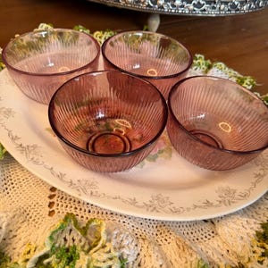May include: Four small, round, translucent purple glass bowls with vertical ribbed detailing along the exterior walls. Each bowl features a circular base with a stamped letter M logo in the center, indicating a consistent design across the set.