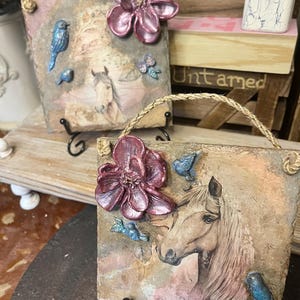 Könnte beinhalten: Zwei quadratische Steinplatten mit einer Pferdekopfillustration, einer großen dreidimensionalen metallisch-magentafarbenen Blume und kleinen blauen Vogelakzenten. Jede Platte hat eine natürliche Steinstruktur, eine Juteschnur und einen schwarzen Metallständer.