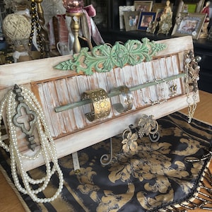 May include: A distressed white wooden jewelry organizer with a green floral accent. The organizer holds bracelets, necklaces, and earrings. A gold and black patterned fabric with fringe is beneath the organizer.