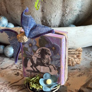 May include: A small, decorative box with a horse illustration, adorned with a blue fabric bow and twine. The box is decorated with a blue flower and moss. The overall theme is rustic and vintage.