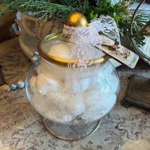 May include: A clear glass jar with a gold rim and lid, filled with fluffy white cotton balls. The lid is topped with a gold sphere and tied with white lace. A decorative tag hangs from the lace.