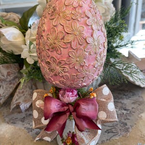 Może przedstawiać: Ozdobne pisanki z różową podstawą i złotymi kwiatowymi zdobieniami. Jajko spoczywa na kompozycji kwiatowej w filiżance do herbaty w stylu vintage, ozdobionej bordową kokardą i wstążką jutową w białe kropki. Urocza dekoracja wielkanocna.