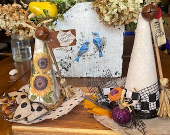 May include: Two cone-shaped decorative ornaments on a wooden board. One features sunflowers, a polka-dot ribbon, and a sign reading Ride Like The Wind. The other is white with a black and white checkered base, a purple pumpkin, and a small tag.