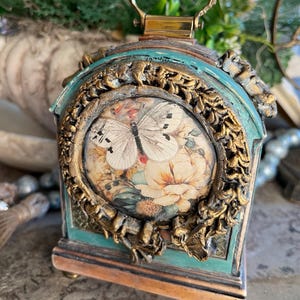 May include: A decorative, antique-style box with a butterfly and floral design. The box features a teal and gold color scheme, with a handle on top. The front of the box displays a circular image of a butterfly and flowers.