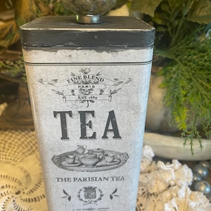 May include: A rectangular white tea tin with a distressed crackle finish, featuring black text that reads FINE BLEND PARIS EST. 1889 TEA THE PARISIAN TEA. The tin has a dark gray lid topped with a round, textured, metallic silver knob.