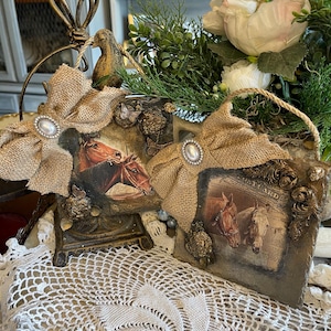 Pode incluir: Duas placas decorativas quadradas com imagens de cavalos, cada uma adornada com um laço de juta e uma pérola central. As placas possuem molduras ornamentadas e texturizadas com detalhes dourados e pequenos elementos florais esculpidos.