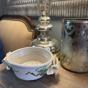May include: A cream-colored ceramic bowl with floral accents. The bowl features a speckled finish and is adorned with pink and green flower and leaf details. A silver mercury glass candlestick and vase are in the background.