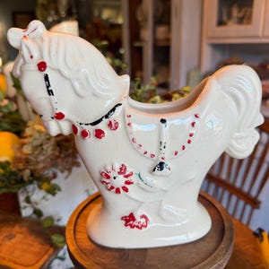 Pode incluir: Um vaso de cerâmica cor de creme em forma de cavalo com acabamento brilhante. O cavalo é decorado com detalhes pintados à mão em vermelho na rédea, sela e um motivo floral na lateral. Possui uma abertura oca nas costas para plantas.