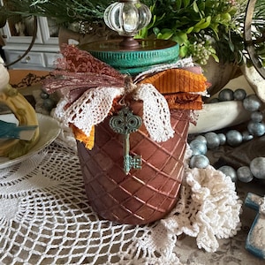 May include: A copper-colored jar with a diamond-patterned texture, topped with a teal lid and a clear glass knob. The jar is adorned with a cluster of orange, brown, and white fabric ribbons and a small, ornate, teal-colored metal key charm.