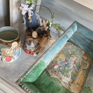 Puede incluir: Colección de artículos decorativos con temática de Pascua. Incluye una bandeja azul y verde con una ilustración de Peter Rabbit, una lata decorada con detalles de mezclilla y encaje, y un pequeño tarro con un diseño de conejito.