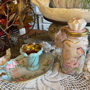 May include: A light blue ceramic dish with pink rose patterns and gold trim, featuring a matching small pedestal cup. Beside it sits a glass jar decorated with floral and bird imagery, topped with a white ceramic knob and a small beige mesh bow.