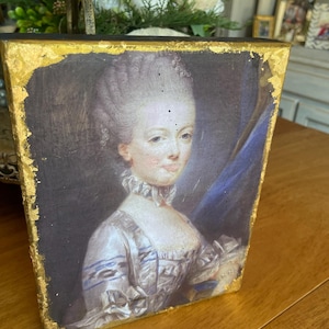 May include: A framed portrait of a woman in a white dress with blue accents. The portrait has a gold leaf border and is set against a dark background. The woman has a powdered wig and a ruffled collar.