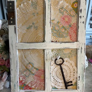 May include: A distressed white framed window, featuring lace doilies and floral fabric within its panes. A decorative black key is attached to the window, creating a vintage aesthetic. The frame has a worn, aged appearance.