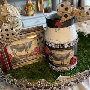 Könnte beinhalten: Eine dekorative Milchkannen aus Metall mit einer schwarz-weiß gepunkteten Schleife und einem gerahmten Kuhdruck. Die Kanne ist weiß mit schwarzen Akzenten und roten floralen Verzierungen. Die Artikel werden auf einem dekorativen Tablett präsentiert.