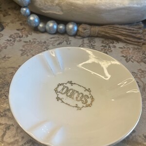 May include: A round white ceramic ashtray featuring three notches along the rim and a gold-colored decorative frame in the center enclosing the word "poros" written in a stylized script font. The edge of the ashtray is finished with a thin gold trim.