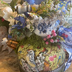 May include: A decorative teapot with a floral design and a bird motif. The teapot is adorned with a variety of flowers, including blue and white blossoms, and is accented with a gingham ribbon. The overall aesthetic is vintage and whimsical.
