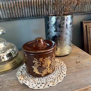 Op de afbeelding: Een kleine, cilindervormige bruine keramieken pot met een bijpassend deksel voorzien van een centrale knop. De buitenkant is versierd met een goudkleurig, sierlijk bloem- en krulmotief, inclusief tekst in cursief schrift.