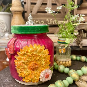 Könnte beinhalten: Dekoratives magentafarbenes Glas mit grünem Samtrand und silbernem Deckel, verziert mit einer großen gelb-orangenen Sonnenblume und einem roten Schmetterling. Im Hintergrund eine kleine Glasvase mit Blumen. Ein dekorativer Gegenstand.