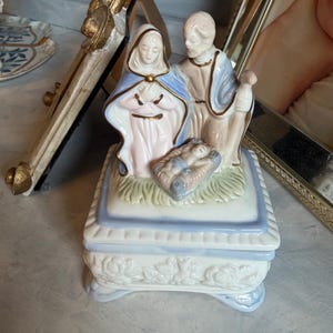 May include: A porcelain music box featuring a nativity scene with a kneeling figure in a blue and white robe, a standing figure in a tan robe, and a baby in a manger. The base is white with blue trim and embossed floral details on the sides.