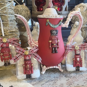 May include: A red teapot with a white lid and handle, decorated with a nutcracker figure. Three nutcracker ornaments in clear containers with sheet music and red and white plaid bows. Christmas decor.