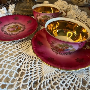 May include: Two porcelain teacups and two matching saucers featuring a deep magenta exterior with gold-toned interiors. Each piece displays a central hand-painted scene of figures in a landscape, accented by gold floral patterns along the scalloped edges.