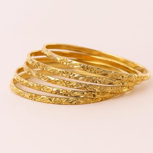 May include: A stack of six gold-colored bangles with a floral pattern. The bangles are stacked on a light pink surface. The bangles have a shiny, reflective surface.