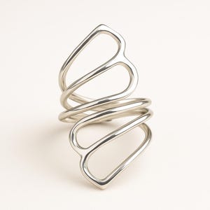 Può includere: Un anello color argento con un design aperto unico. L'anello presenta uno stile a spirale e avvolgente con forme curve e allungate. Il metallo ha una superficie lucida e riflettente, creando un aspetto moderno e artistico.