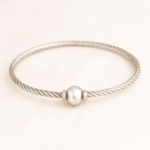 May include: A silver-colored bangle bracelet with a twisted rope design. The bracelet features a central, oval-shaped, pearl-like bead. The bracelet is a simple, elegant piece of jewelry.