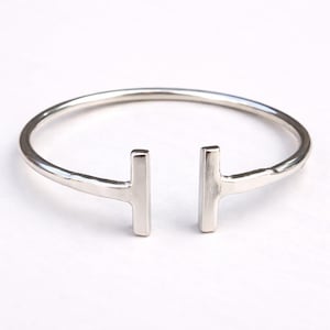 Può includere: Un bracciale aperto color argento dal design minimalista. Il bracciale presenta una fascia sottile e arrotondata che si curva verso due barre verticali rettangolari. Il bracciale è mostrato su uno sfondo bianco.