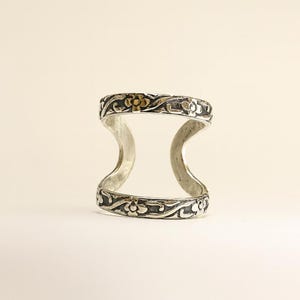 Puede incluir: Anillo de doble dedo plateado con detalles florales y de volutas. El anillo presenta un dise&ntilde;o curvo que conecta dos bandas, con detalles dorados en los motivos florales. El anillo est&aacute; sobre un fondo neutro.