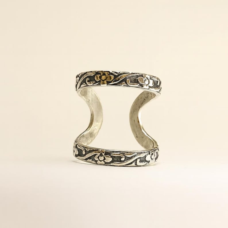 Puede incluir: Anillo de doble dedo plateado con detalles florales y de volutas. El anillo presenta un dise&ntilde;o curvo que conecta dos bandas, con detalles dorados en los motivos florales. El anillo est&aacute; sobre un fondo neutro.