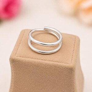 Può includere: Un anello color argento lucido caratterizzato da un design bypass con due fasce parallele che si sovrappongono leggermente. L'anello presenta una finitura metallica liscia e riflettente e una struttura semplice con bordi arrotondati.