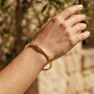 May include: A gold-coloured bangle bracelet featuring square-cut clear gemstones set into the top surface. The bracelet is shown on a person's wrist and arm, with a blurred background of foliage and a stone wall.