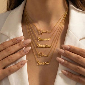 Puede incluir: Collares dorados con nombres: Ophelia, Summer, Madeline, Olivia y Simona. Los collares los lleva una persona con una blazer blanca. Los nombres están en diferentes fuentes.