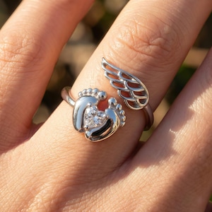 Pode incluir: Um anel prateado com pegadas de bebé a abraçar uma gema em forma de coração. Um design de asa aberta está na lateral. O anel é usado num dedo, com um fundo desfocado.