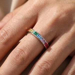 May include: A silver ring with a band of colorful gemstones in a rainbow pattern. The gemstones are square-cut and arranged in a spectrum of colors, including red, orange, yellow, green, blue, and purple. The ring is worn on a finger.