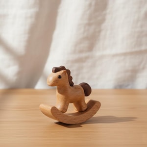 Könnte beinhalten: Ein kleines Schaukelpferd aus Holz. Das Pferd ist hellbraun mit einer dunkelbraunen Mähne und Schweif. Es ist auf gebogenen Kufen montiert. Das Spielzeug ist für Kinder konzipiert und aus Naturholz gefertigt. Ca. 15 cm lang.