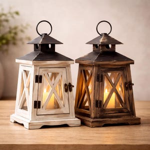 Może przedstawiać: Dwie dekoracyjne drewniane latarnie. Jedna pomalowana na biało z czarnymi akcentami, druga bejcowana na brązowo. Obie mają czarny metalowy daszek i uchwyt. Każda latarnia ma szklany panel i zapaloną świecę w środku.