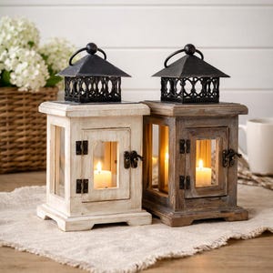 Puede incluir: Dos faroles decorativos de madera, uno blanco y otro marrón, cada uno con una parte superior y un asa de metal negro. Los faroles tienen paneles de vidrio y contienen velas encendidas. Se exhiben sobre una tela de color neutro.