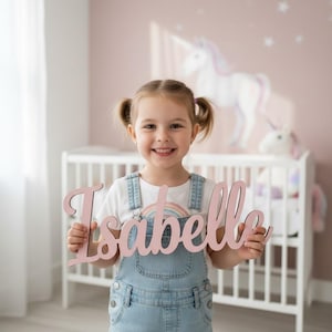 以下が含まれることがあります： 若い女の子が、筆記体で「Isabelle」と書かれたピンク色の木製サインを持っています。サインは、ユニコーンの壁のデカールとベビーベッドを含む、ユニコーンをテーマにした保育園の中にあります。