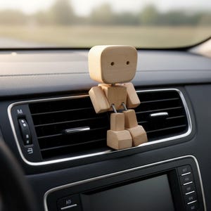 May include: A wooden car air vent decoration shaped like a small person. The figure has a square head with two eye holes and articulated arms and legs. It is light brown in color and sits on a black car dashboard.