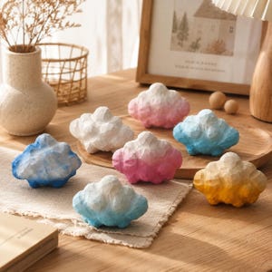 Puede incluir: Una colección de siete pequeños objetos decorativos con forma de nube y superficie tallada y texturizada. Cada pieza presenta un degradado de color único, incluyendo tonos de blanco, rosa, azul y amarillo, dispuestos sobre una superficie de madera.