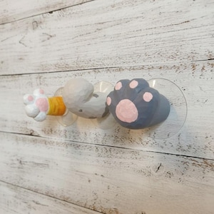 Puede incluir: Un juego de tres ganchos decorativos con ventosa en forma de patas de gato. Las patas están pintadas en blanco, gris y azul con detalles en rosa. Los ganchos están fijados a una pared de madera blanca.
