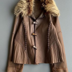 May include: A brown leather jacket with a faux fur collar. The jacket features decorative stitching and three toggle closures. The sleeves are long, and the jacket has a flared shape.