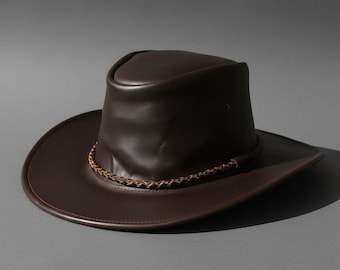 Peut inclure: Chapeau en cuir marron foncé avec un large bord et une bande tressée autour de la couronne. Le chapeau a un design de chapeau de cowboy classique, avec un bord légèrement incurvé et une couronne arrondie. En cuir lisse.
